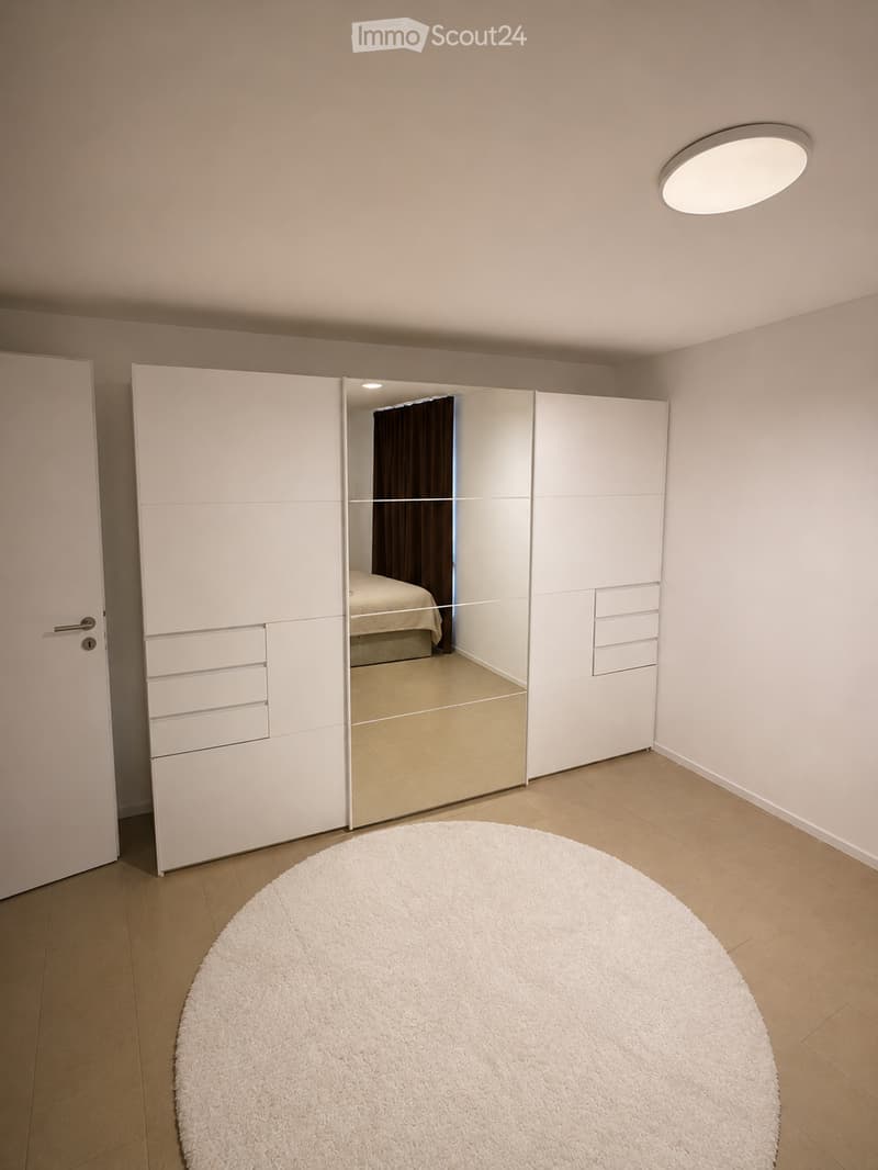Empty room with light-colored walls, wooden flooring, a large white sliding wardrobe with mirrored doors, and a round white rug in the center.