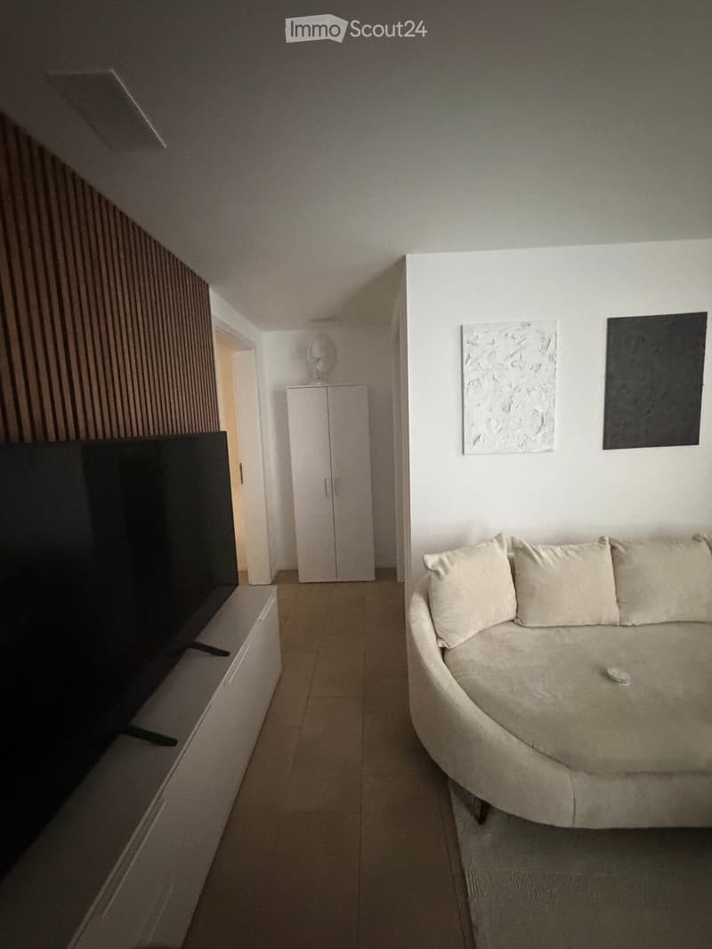 Living room with a beige sofa, a white cabinet, a large screen on a stand, and a small table with a decorative item on top.