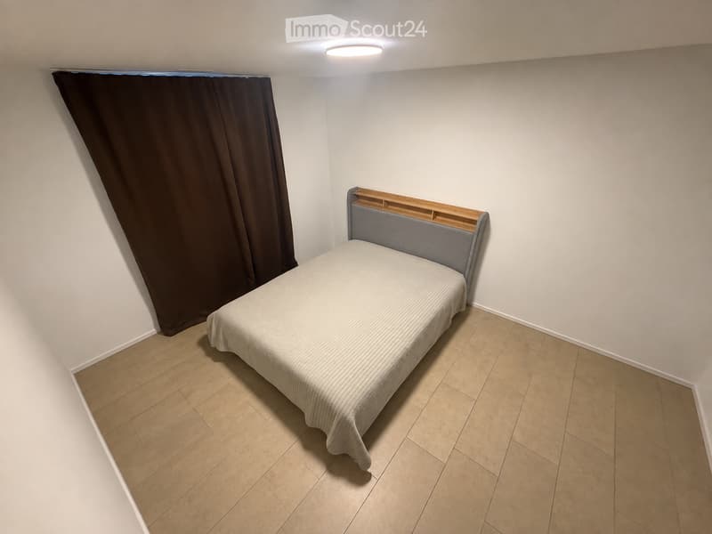 Minimalist bedroom with a single bed, grey headboard, and light-colored bedding, set against white walls and light wooden flooring.