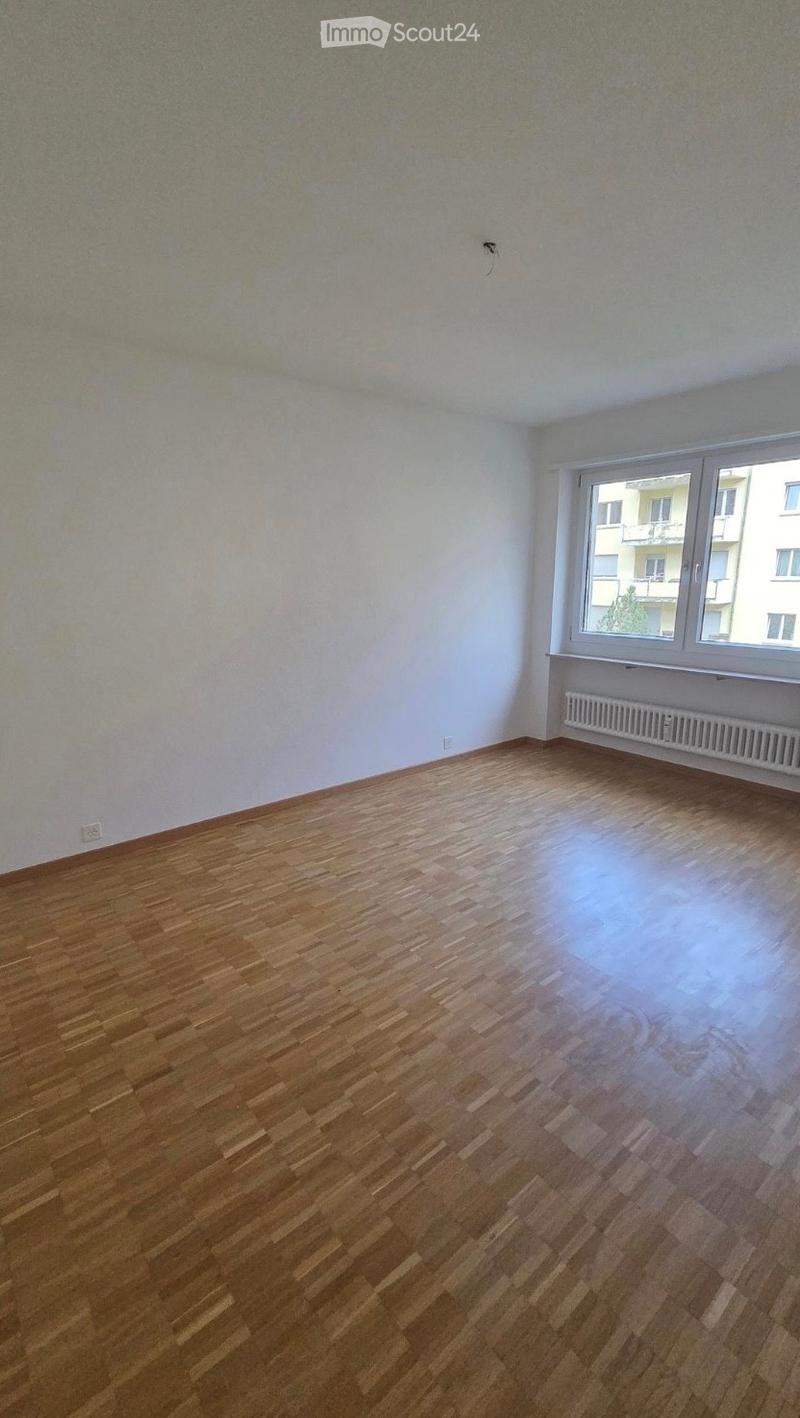 Empty room with wooden flooring and white walls, featuring a large window with a view of neighboring buildings and a radiator below the window.