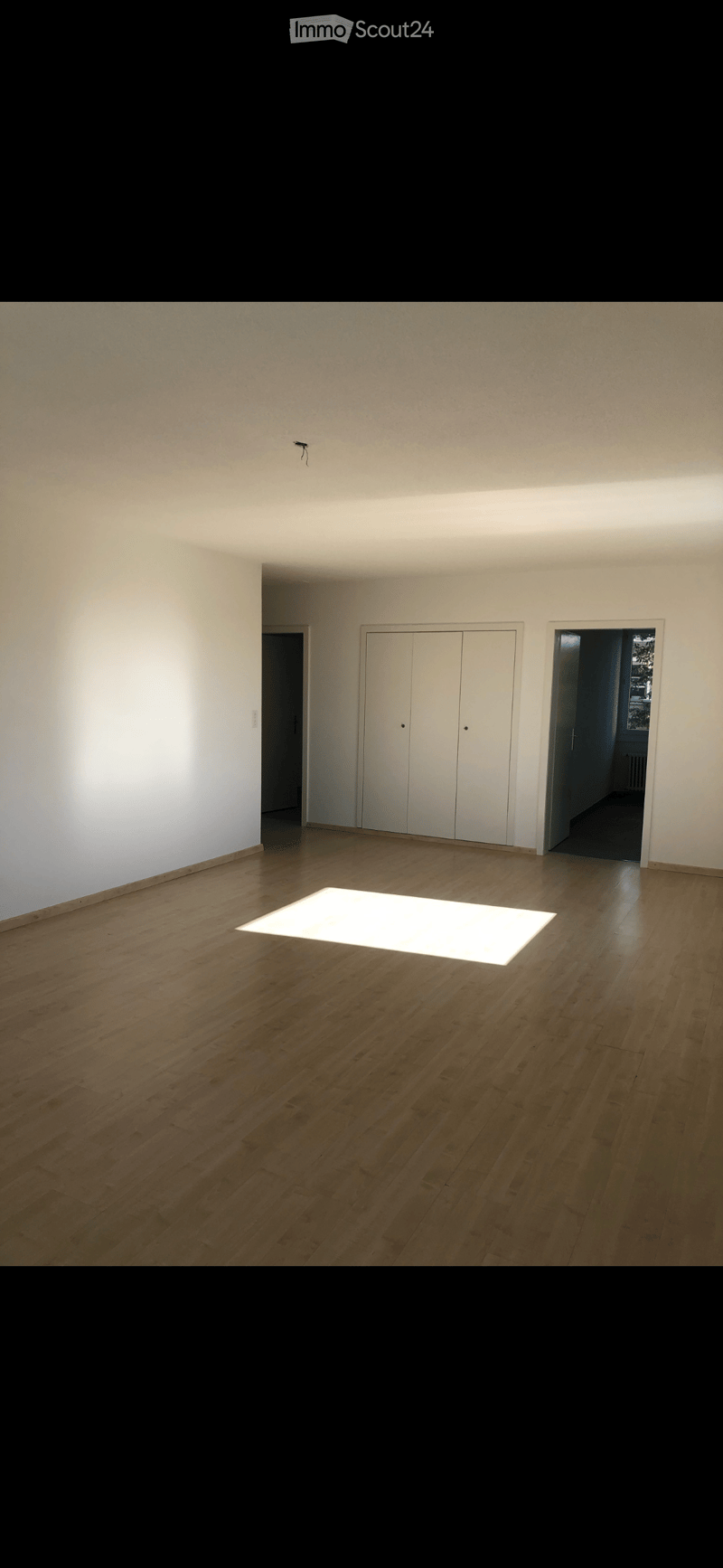 Empty room with wooden flooring, white walls, and a large illuminated square in the center. There are two white closet doors and an open doorway leading to another space.