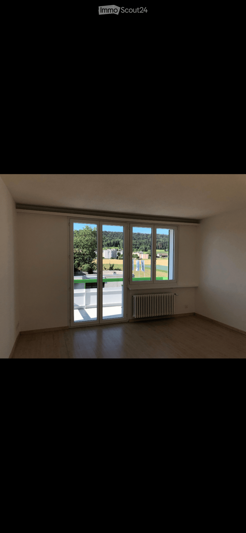 Empty room with wooden flooring, large sliding glass doors leading to a balcony, radiator, and a view of greenery and buildings.