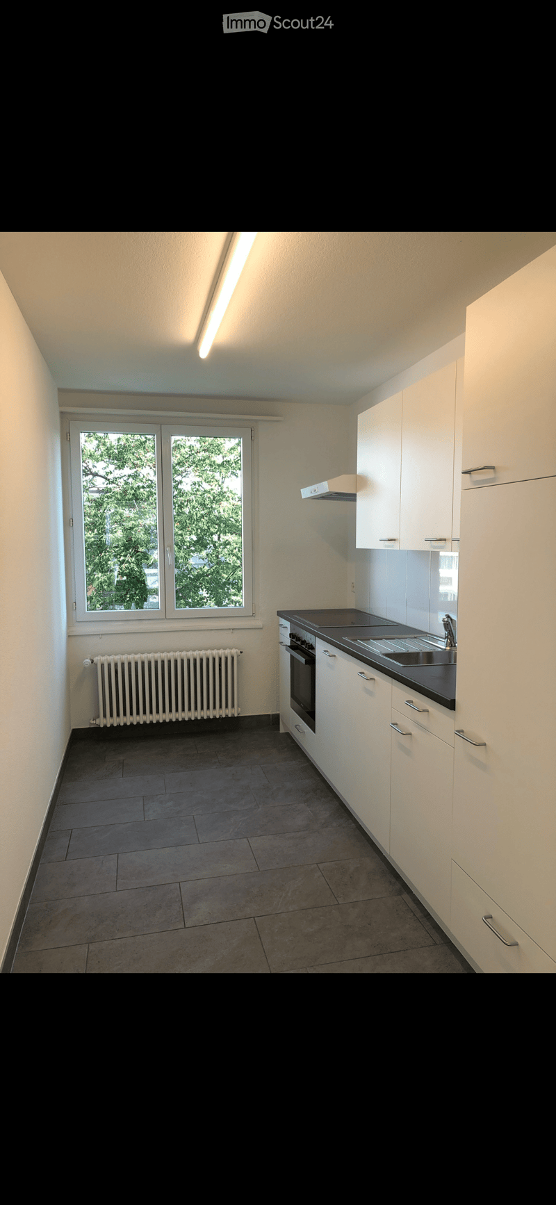 Empty kitchen with white cabinets, dark countertops, a sink, and a window overlooking greenery. The floor is tiled, and there is a radiator below the window.