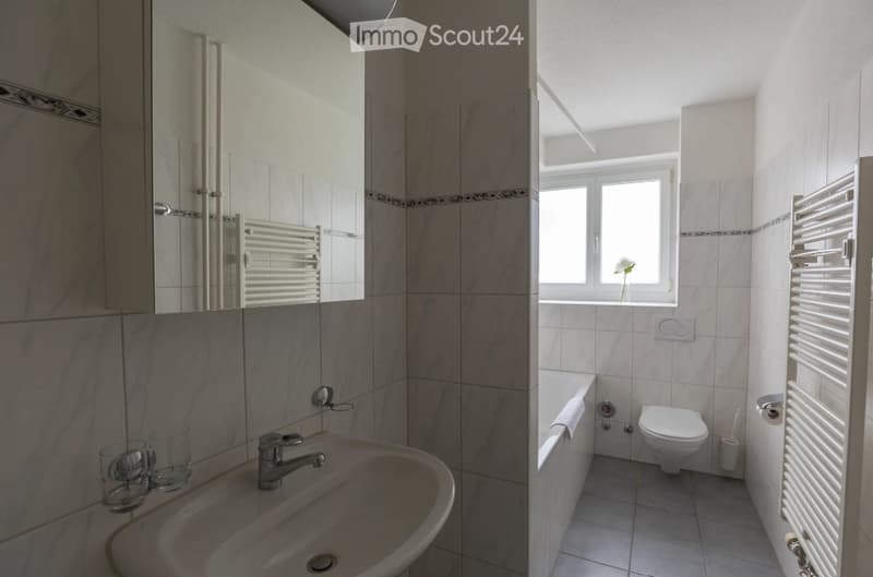 Modern bathroom with a sink, mirror, radiator, bathtub, toilet, and a window with a flower in the corner.