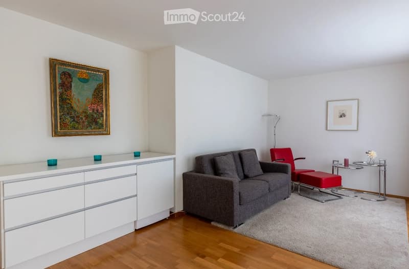 Modern living room with a grey sofa, red armchair, small round table, white cabinet, and artwork on the walls.