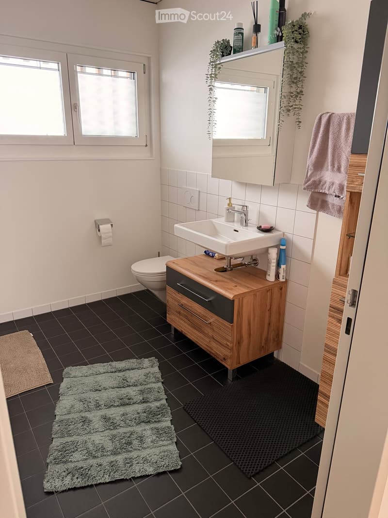 Modern bathroom with dark tiled floor, white walls, sink with wooden cabinet, toilet, mirror, and towels.