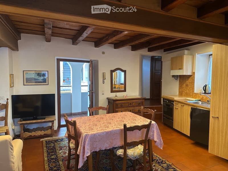 A cozy living and dining area with a wooden ceiling, a dining table with chairs, a TV, a kitchen area with cabinets, sink, oven, and refrigerator, and a small bathroom visible in the background.