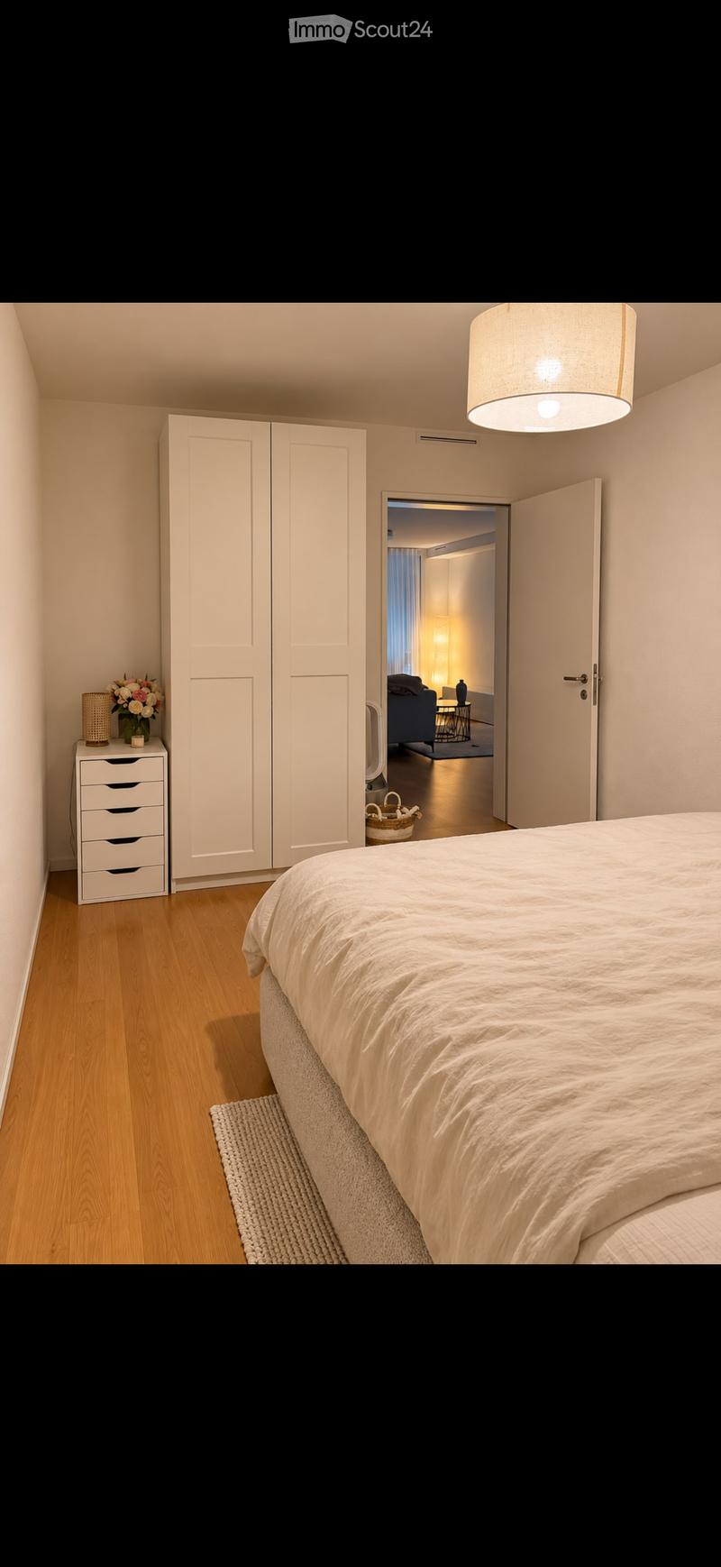 Modern bedroom with a large bed, white wardrobe, nightstand with a flower vase and lamp, and an open door leading to a living area with a sofa and coffee table.