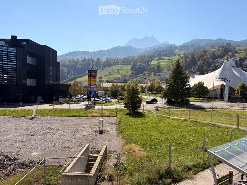 View of a park area with grassy patches, gravel pathways, a metal fence, and a modern building in the background, with mountains visible in the distance.