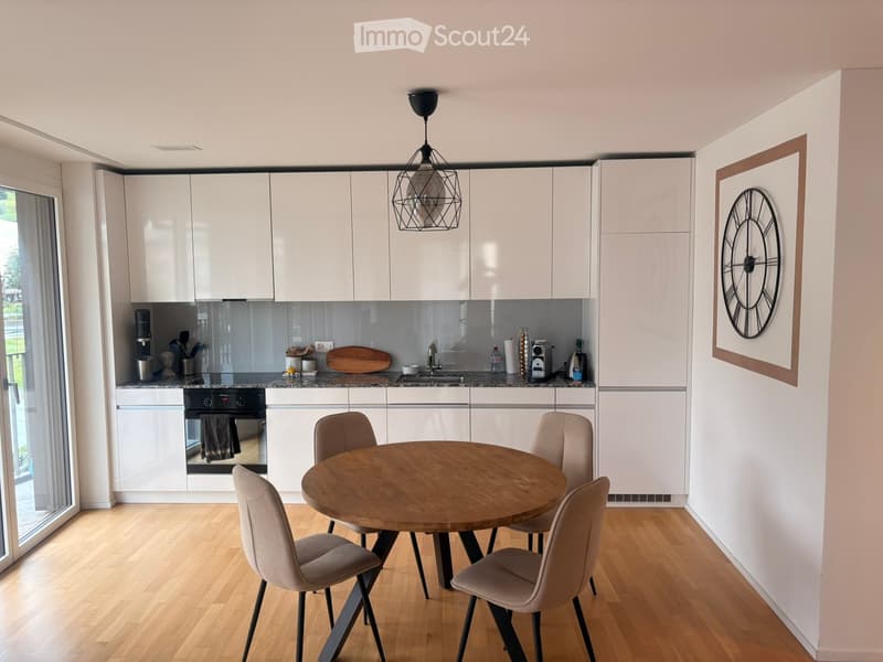 Modern kitchen with white cabinets, a round wooden dining table with four chairs, a hanging pendant light, and a stainless steel oven.