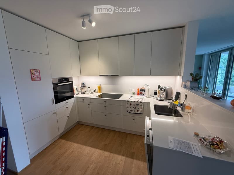 Modern kitchen with white cabinets, stainless steel appliances, and a light wooden floor. Features an oven, sink, coffee maker, and various small appliances on the countertops.