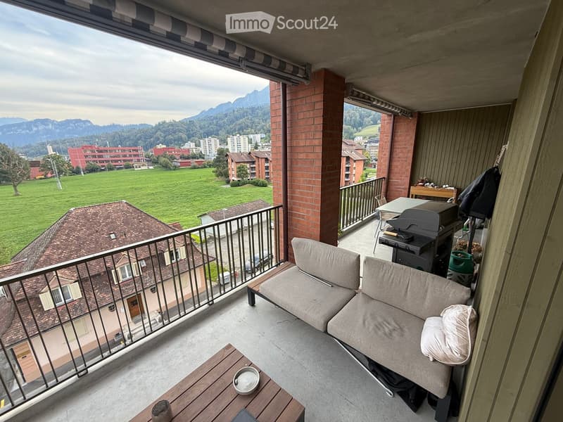 Balcony with a view of a green field and mountains, furnished with a sofa, small table, grill, and chairs.