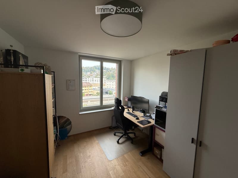 A modern office space with a wooden floor, large window offering a city view, a desk with a computer setup, a comfortable office chair, and built-in storage cabinets.