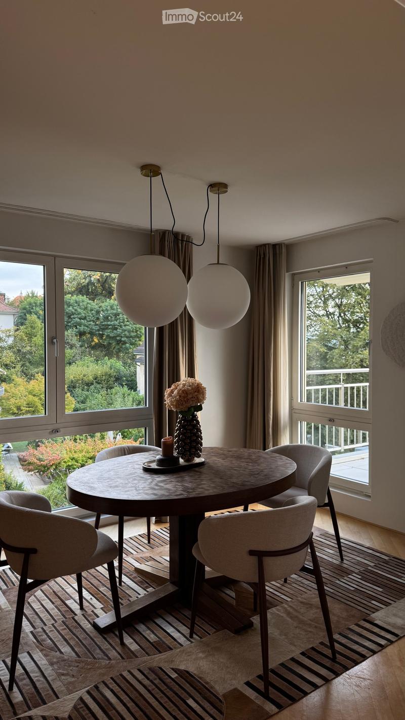 Modern dining room with a round wooden table, four chairs, a vase with flowers, and pendant lights. Large windows offer a view of the garden.