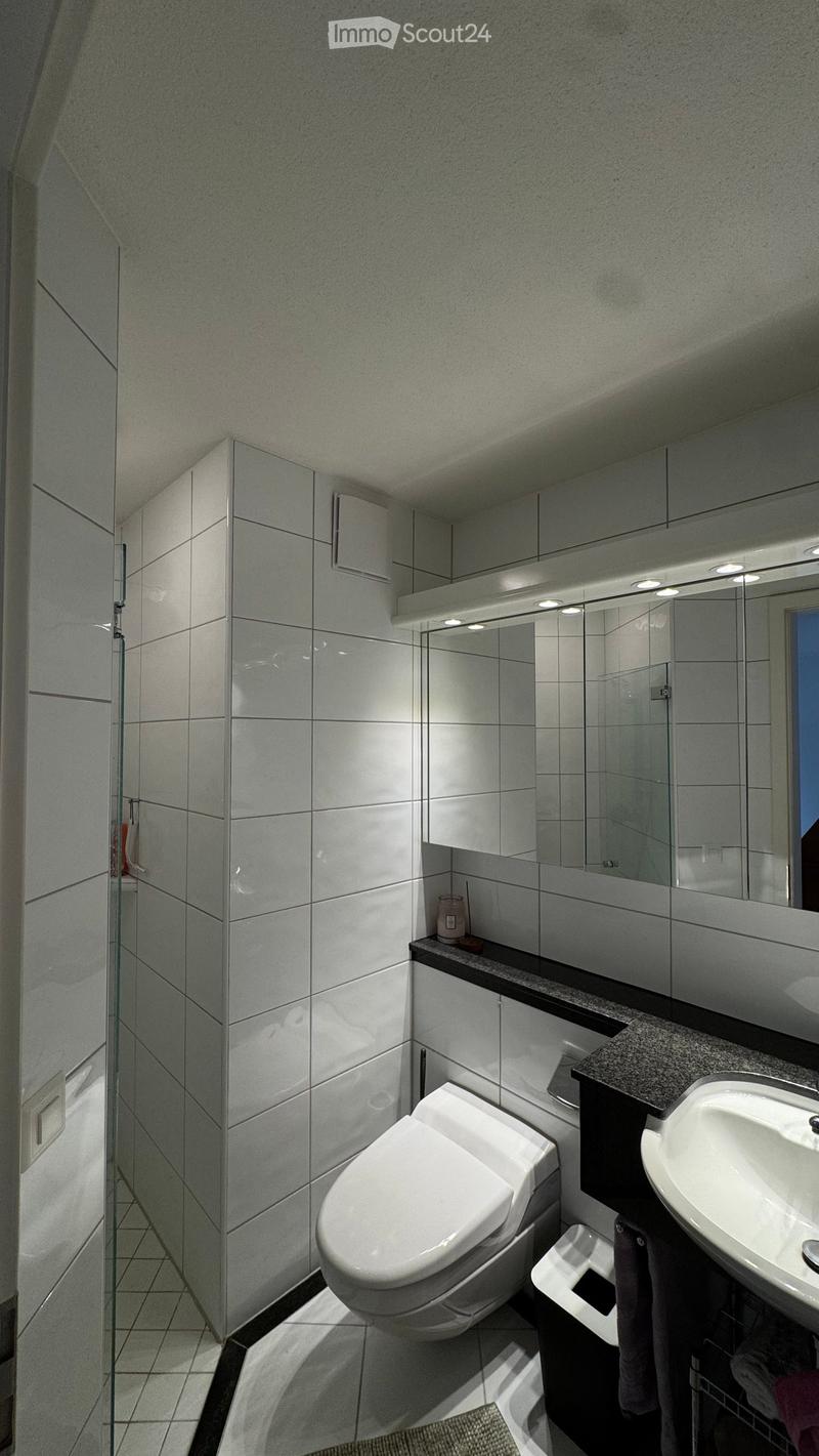 Modern bathroom with white tiles, a toilet, a sink, a large mirror with built-in lighting, a glass-enclosed shower, and a small cabinet with a towel visible.