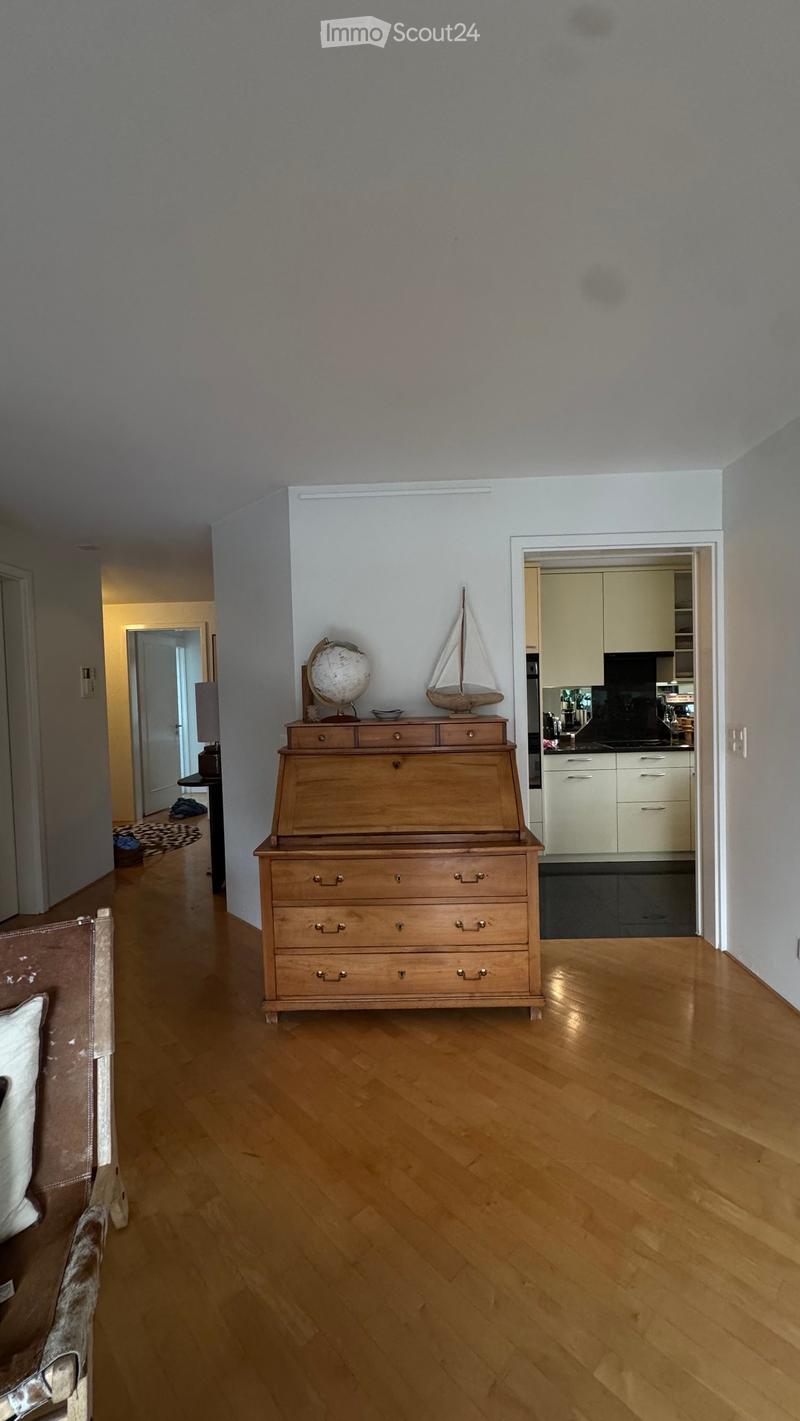 A well-lit hallway with wooden flooring, a chest of drawers with a globe and model sailboat on top, and a glimpse of a modern kitchen with white cabinets and stainless steel appliances.