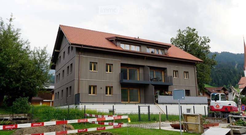 Modern two-story house with a red-tiled roof, large windows, and a balcony on the upper floor. The house is surrounded by greenery and has a fenced yard with construction barriers.