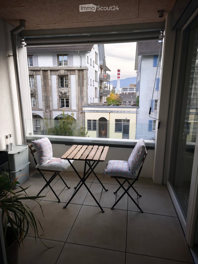 Balcony with a small wooden table and two matching chairs, overlooking a cityscape with buildings and a distant view of mountains.