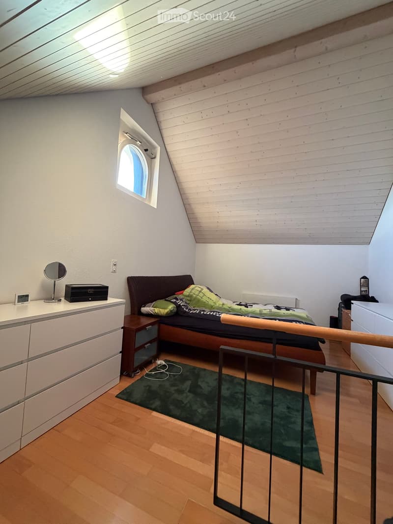 Cozy attic bedroom with wooden slanted ceiling, bed with dark headboard, nightstand, mirror, and small rug.