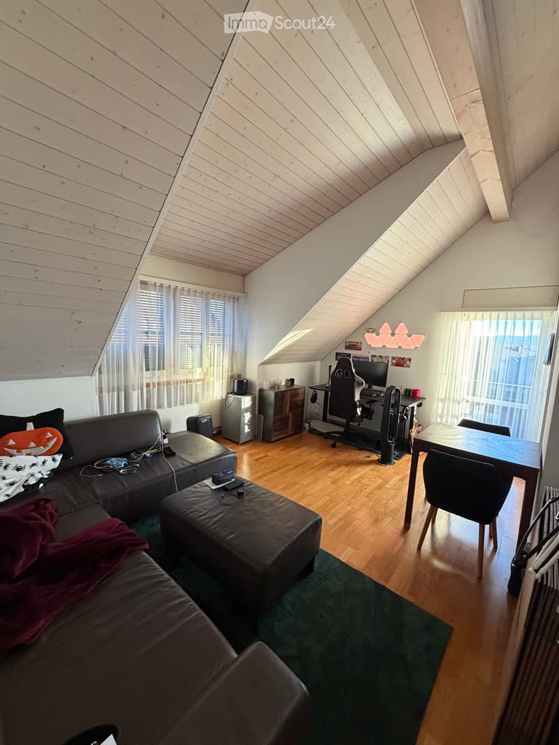 A cozy attic room with wooden slanted ceilings, a black sectional sofa, an ottoman, a small table, a computer desk with a chair, and a dining table with chairs. The room features large windows with white curtains and a green rug.