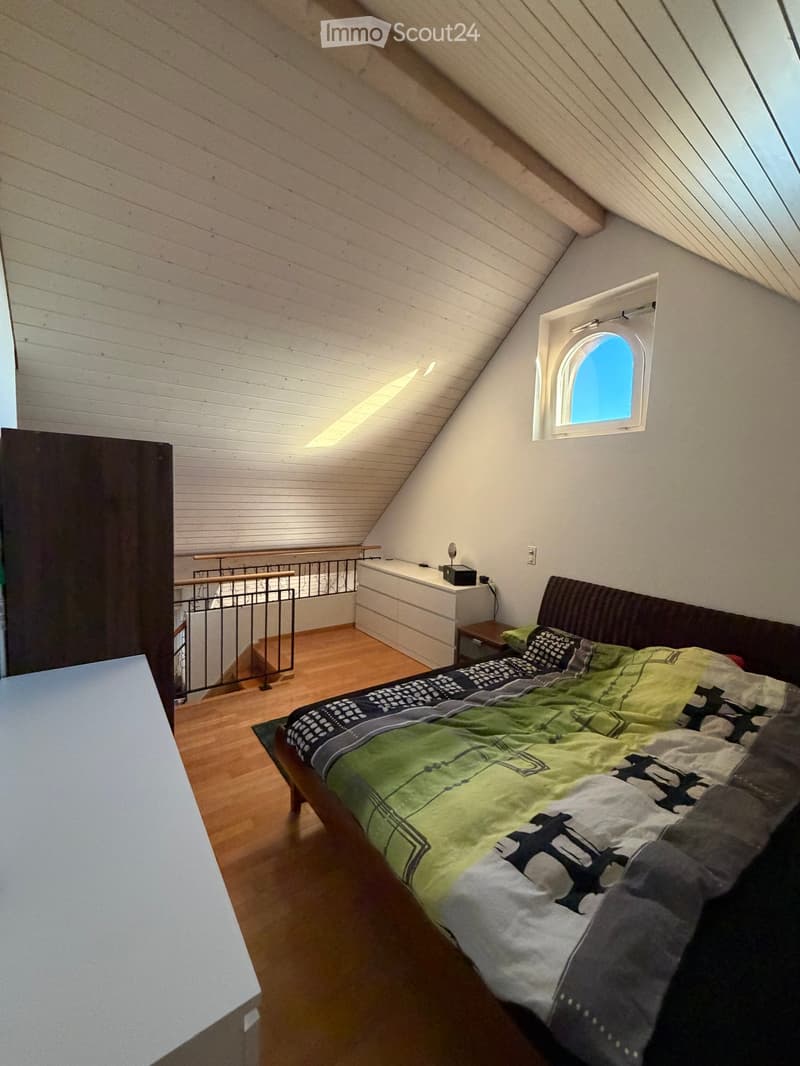 Cozy attic bedroom with wooden floors, slanted wooden ceiling, skylight window, bed with green and grey bedding, nightstand, small table, and chair.