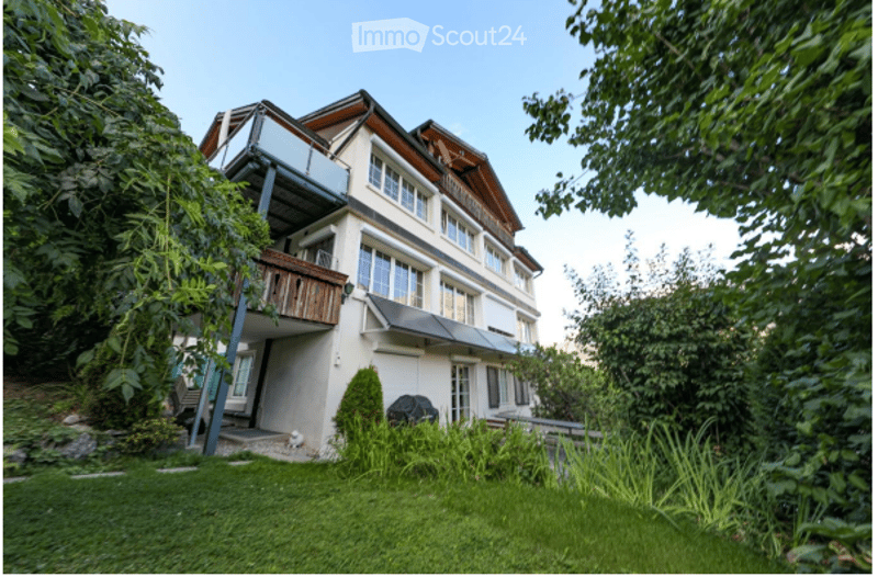 Modern two-story house with white walls, large windows, and a balcony on the upper level. The house is surrounded by a well-maintained garden with green grass and trees.