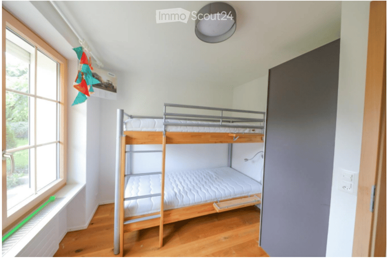 A small bedroom with a wooden bunk bed, white walls, wooden flooring, and a window with a view of greenery.