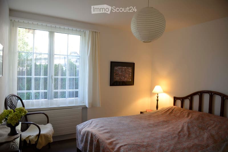 Cozy bedroom with a double bed, a wooden chair, a small table with a vase of flowers, a bedside lamp, and a large window with white curtains. A decorative picture frame hangs on the wall, and a large paper lantern hangs from the ceiling.
