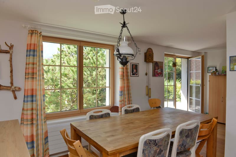 Bright kitchen with large windows, wooden dining table surrounded by chairs, cabinet, picture frames, and a hanging light fixture.