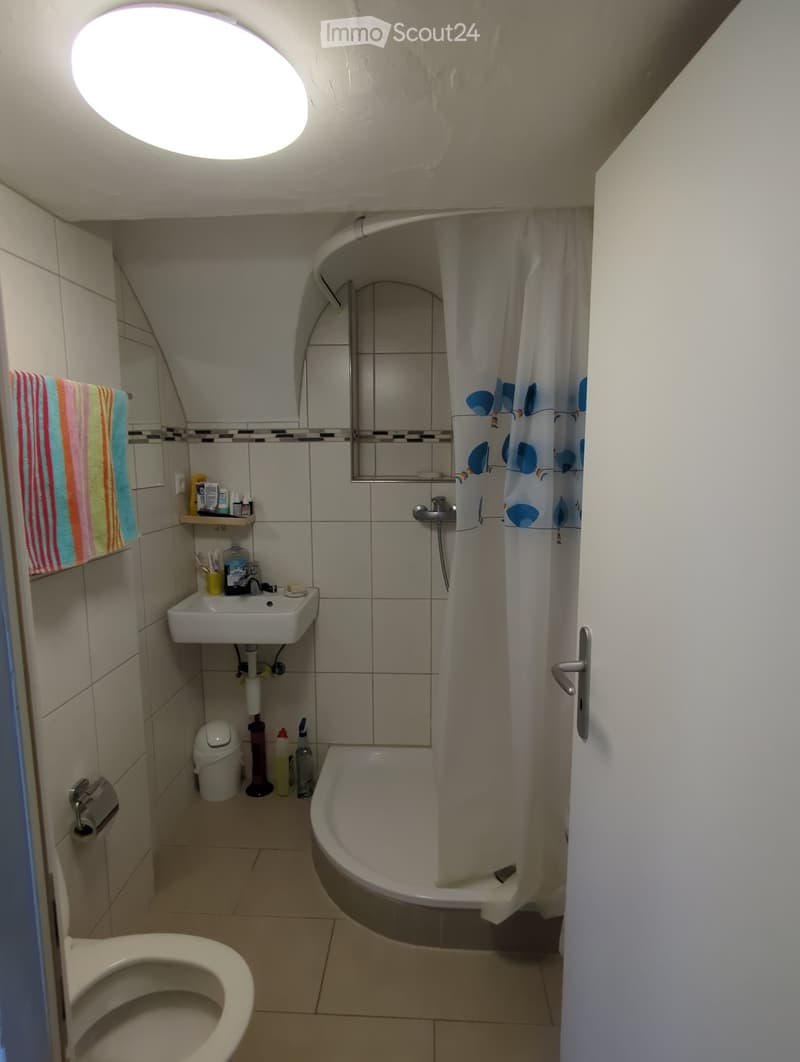 Small bathroom with a toilet, sink, bathtub, and mirror. Features a colorful towel, a shower curtain, and various toiletries on the sink.