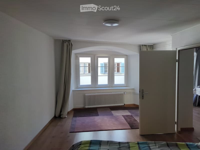 Empty bedroom with wooden flooring, large window with curtains, radiator below, and a partially visible bed with a plaid blanket in the foreground.