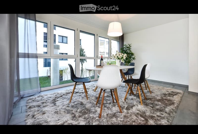 Modern dining area with large windows offering a view, a round white dining table surrounded by black and white chairs, a fluffy white rug, and a flower vase on the table.