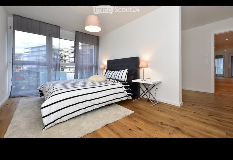 Modern bedroom with a large bed, nightstand, table lamp, and large windows with curtains. The room features wooden flooring and a white rug.