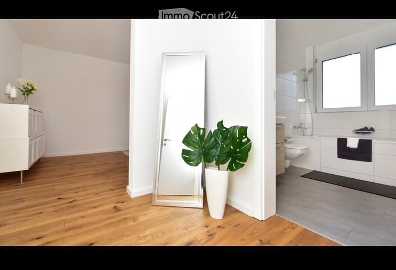 Modern, minimalist room with wooden flooring, large mirror, potted plant, and adjacent bathroom with bathtub, toilet, and shower.