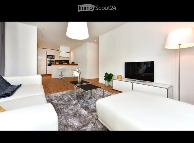 Modern living room with white sofa, coffee table, two stools, television, floor lamp, and two small plants. The kitchen area features white cabinets, an oven, microwave, and refrigerator.