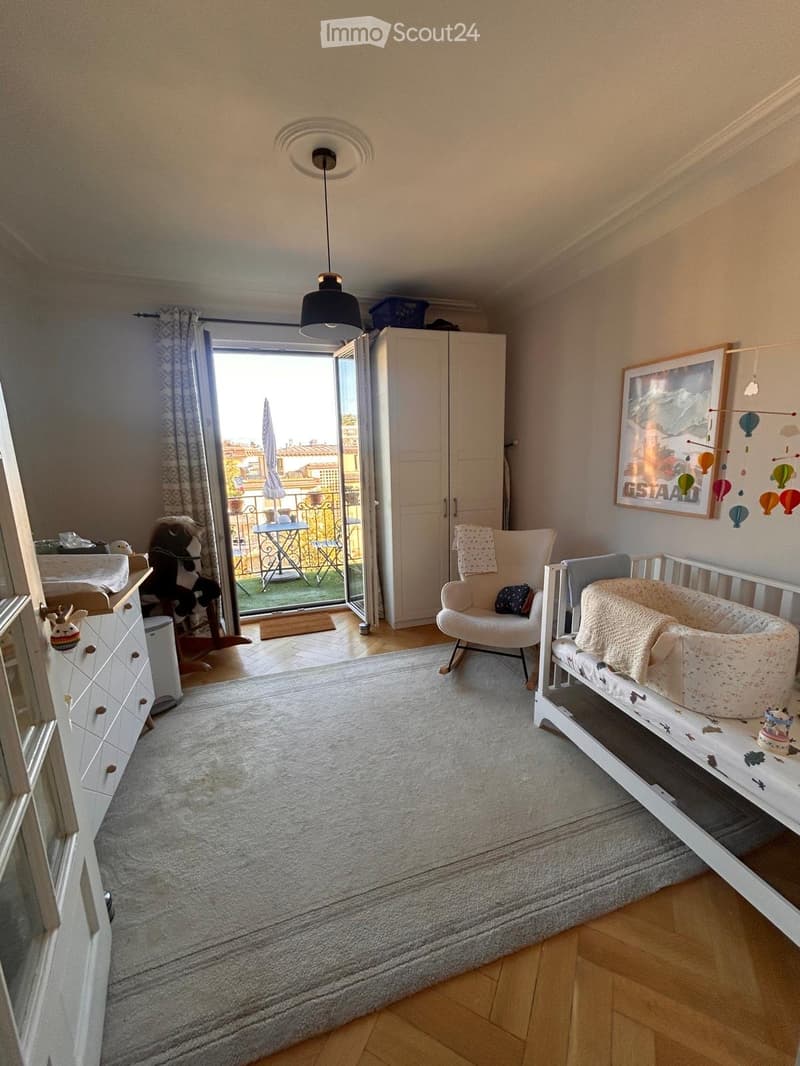Brightly lit bedroom with a crib, rocking chair, cabinet, and toys. Large window opens to a balcony with outdoor furniture.