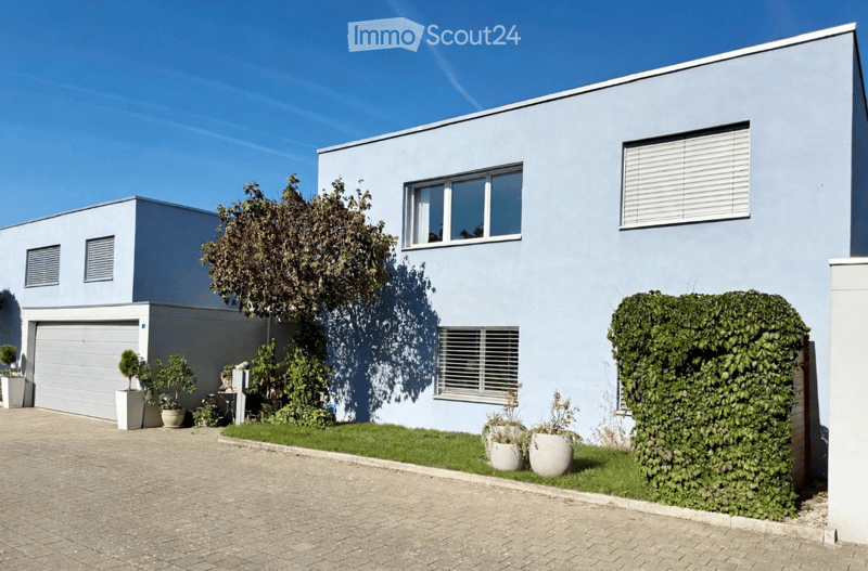 Modern two-story house with light blue exterior, garage, and small garden area with potted plants.