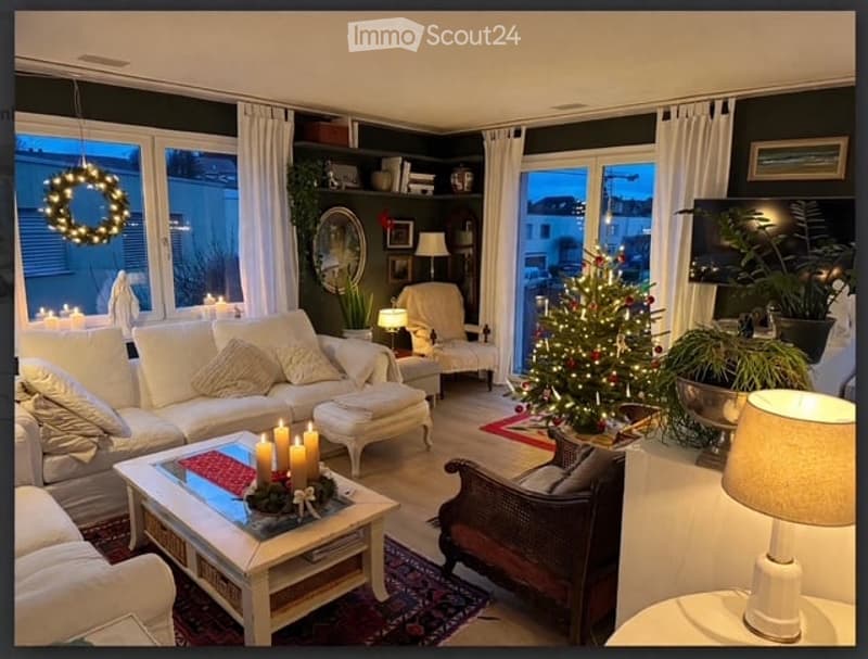 Cozy living room with large windows, white sofa, coffee table with candles, armchair, ottoman, floor lamp, table lamp, television, Christmas tree, wreath, mirror, and potted plant.