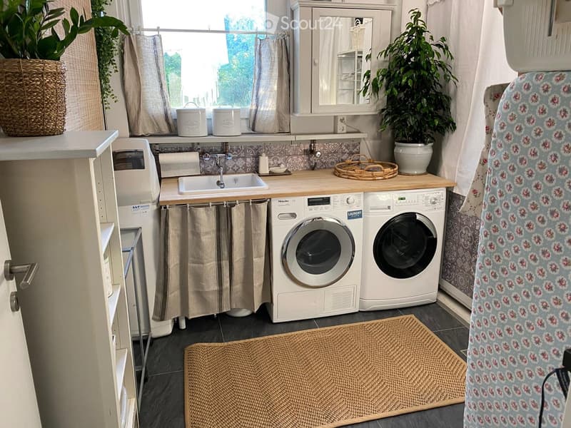 Well-lit laundry room with a sink, washing machine, dryer, and a mirror above the sink. Features wicker baskets, plants, and a decorative towel rack.
