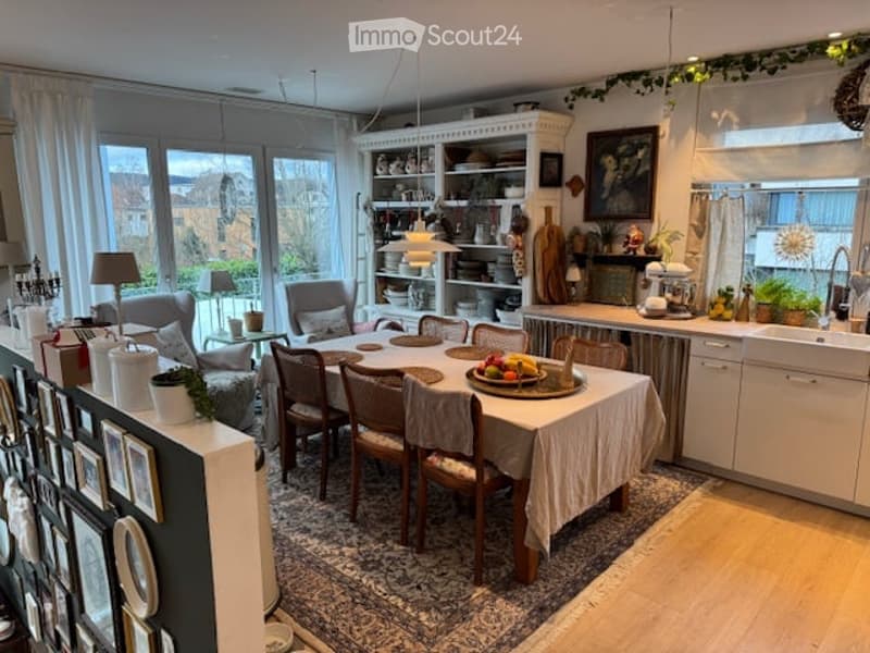 Open-plan living and dining area with a dining table, chairs, sofa, bookshelf, kitchen cabinets, sink, and various decorative items.