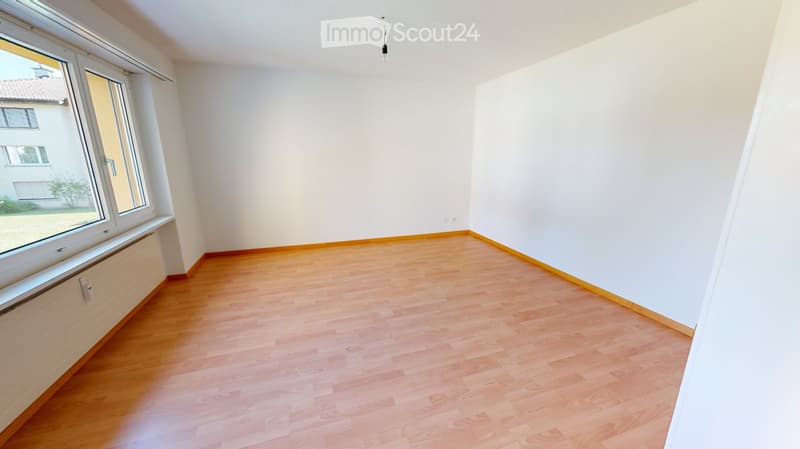 Empty room with light wooden flooring, white walls, large window, radiator, and visible electrical outlet.