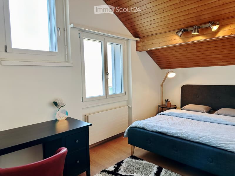 Cozy bedroom with a double bed, wooden desk, red chair, and a vase with white flowers on the desk. The room features wooden flooring, white walls, and large windows with white frames.