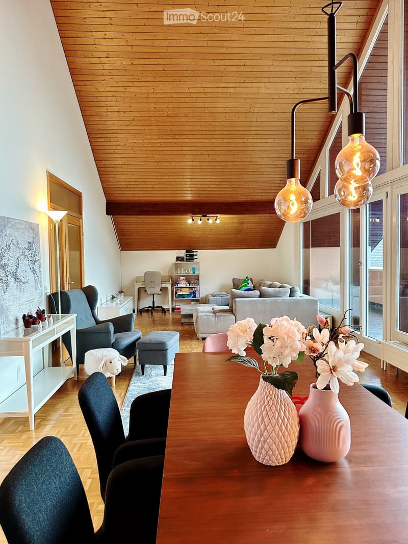 Bright, modern living room with wooden floors and a slanted wooden ceiling. Features a sofa, chairs, a coffee table with vases, a small shelf, and a hanging lamp with multiple bulbs.