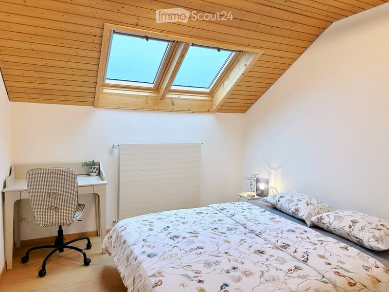 Cozy attic bedroom with wooden ceiling, skylight windows, a double bed with floral bedding, a desk with a chair, and a small side table with a lamp and plant.