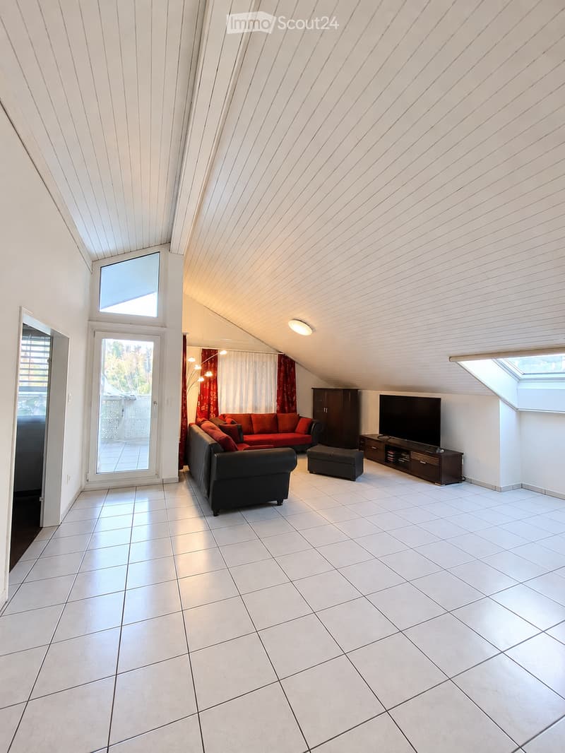 Modern living room with white tiled flooring, slanted ceiling, large windows, and a balcony. Features a black sofa, red ottoman, TV, and wooden cabinet.