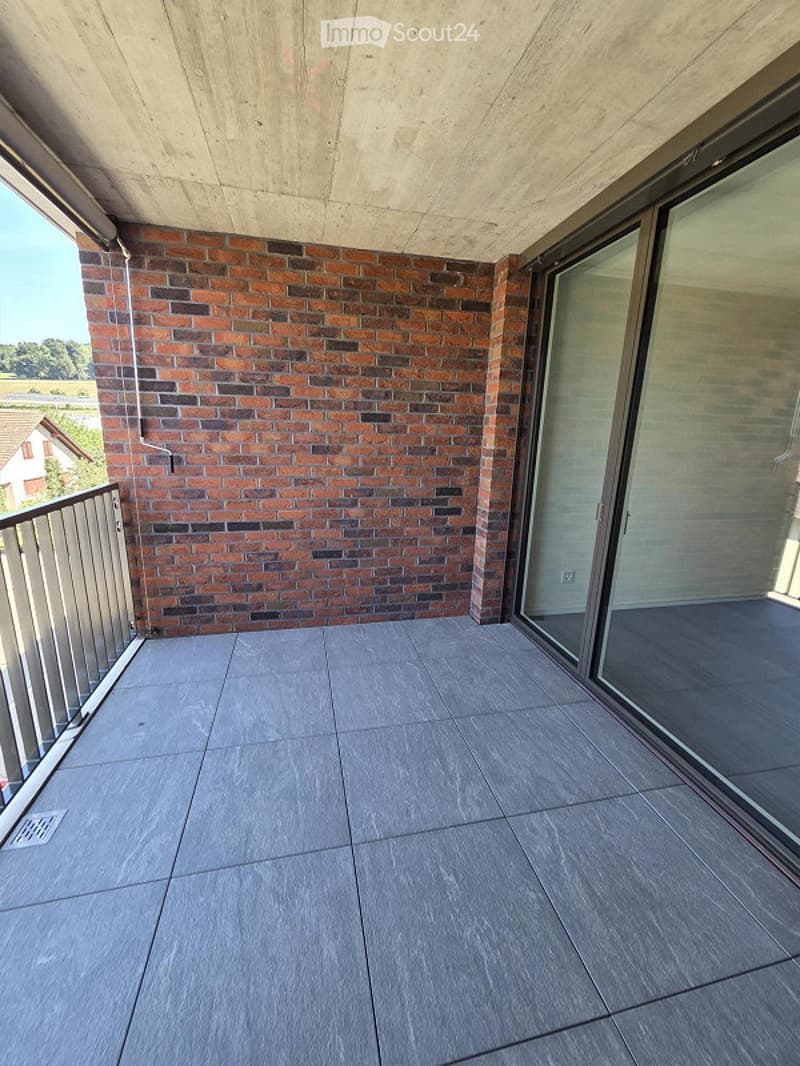 Modern balcony with grey tiled flooring, metal railing, and large glass sliding doors leading to an interior space. The balcony features a brick wall backdrop and offers a scenic view of a rural landscape.