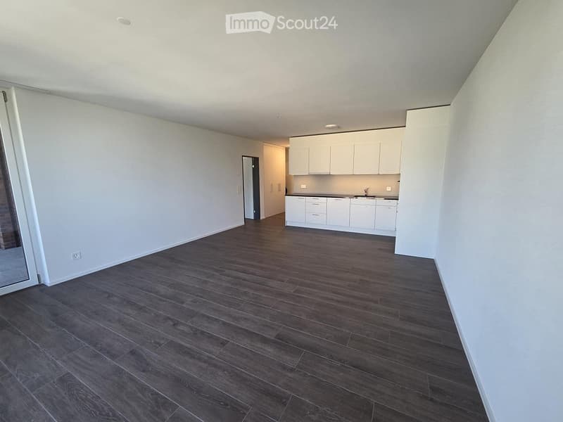 Empty room with dark wooden flooring and white walls, featuring a small white kitchen area in the background.