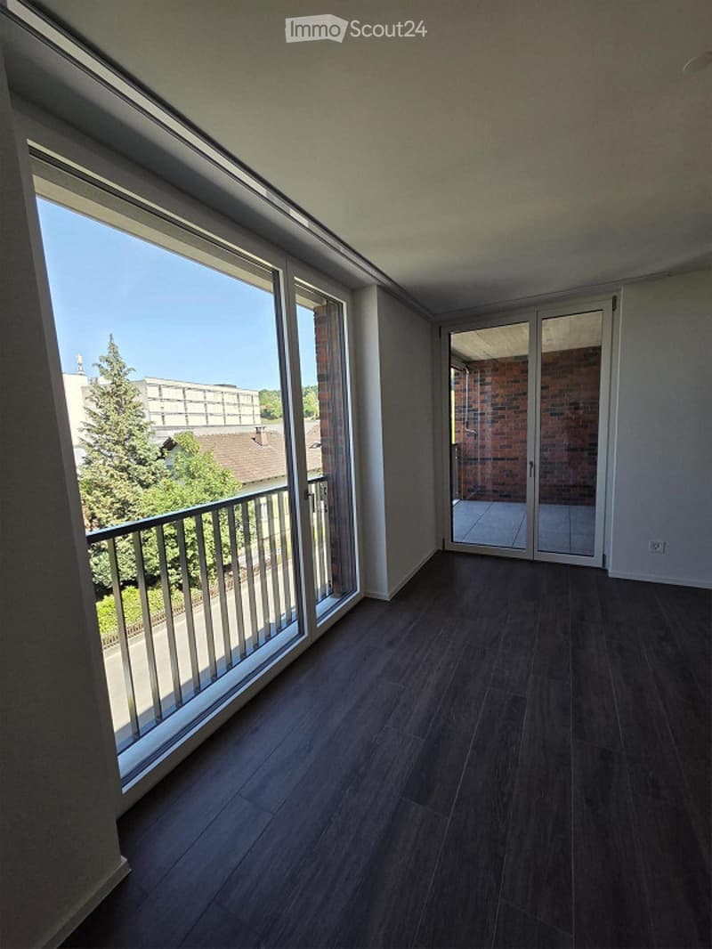 Empty room with dark wooden flooring, large windows, and a sliding glass door leading to a balcony with a view of trees and buildings.