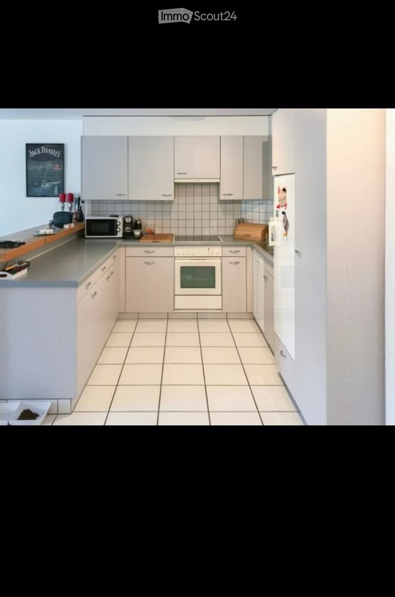 Modern kitchen with white cabinets, tiled floor, and stainless steel appliances including a microwave, oven, and refrigerator. Countertop features a cutting board and various small items.