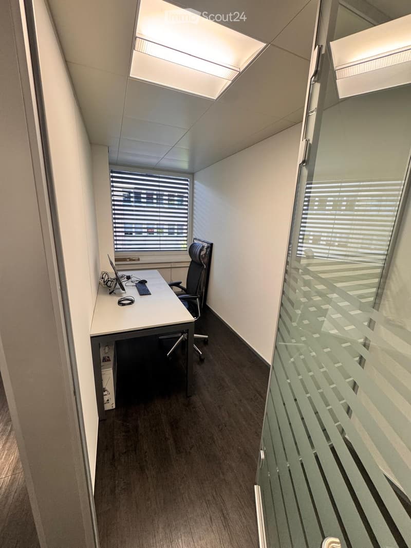 Modern office space with a desk, chair, laptop, and keyboard. Large windows with blinds and a glass partition.